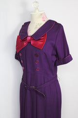 Women's vintage 1960's Mode O'Day label short sleeve knee length purple wool blend dress with peter pan collar, satin bow, matching belt, and decorative buttons.