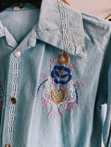 Women's or men's vintage 1970's long sleeve light wash denim shirt with popper buttons, pockets, belt loops, and floral embroidery throughout.