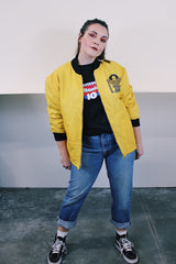 Women's or men's vintage 1970's long sleeve yellow nylon zip up bomber jacket with black knit trim and black graphic on left chest.