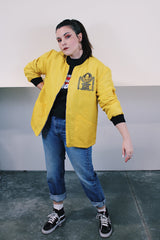 Women's or men's vintage 1970's long sleeve yellow nylon zip up bomber jacket with black knit trim and black graphic on left chest.
