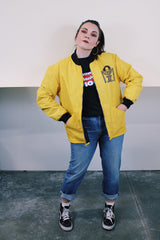 Women's or men's vintage 1970's long sleeve yellow nylon zip up bomber jacket with black knit trim and black graphic on left chest.