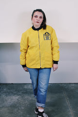 Women's or men's vintage 1970's long sleeve yellow nylon zip up bomber jacket with black knit trim and black graphic on left chest.