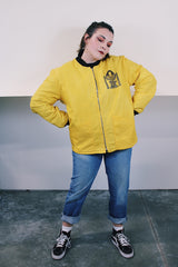 Women's or men's vintage 1970's long sleeve yellow nylon zip up bomber jacket with black knit trim and black graphic on left chest.