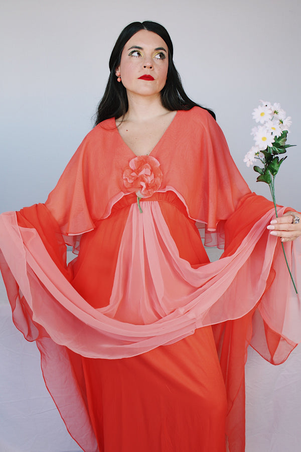 Women's vintage 1980's maxi dress in peach coral color with a V shaped neckline, cape like sleeves, and flower on chest