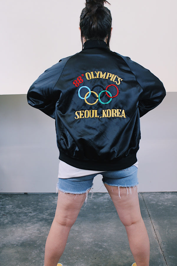 Women's or men's vintage 1988 long sleeve black shiny stain polyester bomber jacket with side pockets. Has 88' Olympics Seoul Korea embroidered on the back with Olympics logo.