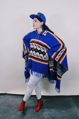 Women's or men's vintage 1970's blue acrylic material poncho with fringe trim and half zipper closure at neck. White. black, brown, and red striped print. 